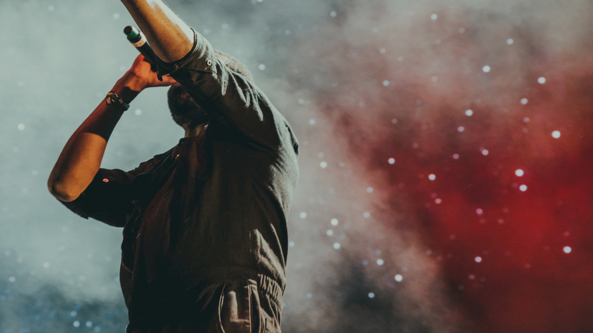 Sångare på scen sjunger i mikrofon med ena handen upphöjd. Rökig bakgrund i grå och röd ton skapar en dramatisk stämning.