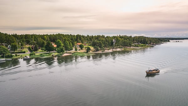Flygbild över Kristinehamns skärgård under en solnedgång. En ensam båt glider fram över spegelblankt vatten. Längst bort i bort i bild syns strandkanten, mede båtbryggor och den angränsande skogen.